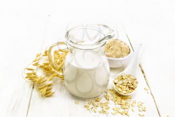 Oat milk in a jug, flour in bowl, oatmeal in spoon, stalks oaten on white wooden board background