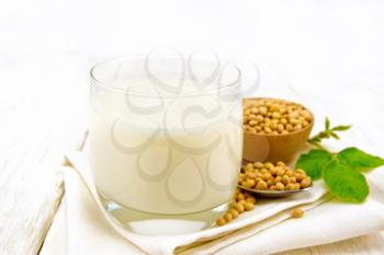 Soy milk in a glass, green leaf, soybeans in a spoon and bowl on a napkin against the background of a light wooden board