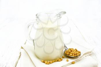 Soy milk in a jug, soybeans in a spoon on a napkin against the background of white wooden board