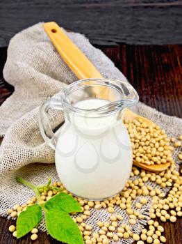Soy milk in a jug, green leaf, soybeans in a spoon on a napkin of burlap on the background of a dark wooden board