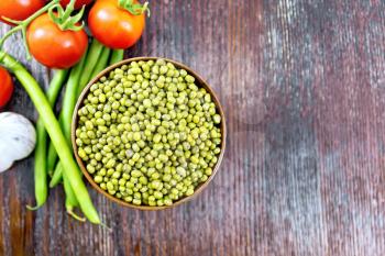 Green lentils mung in a bowl, pods of beans and red tomatoes on a wooden board background from above