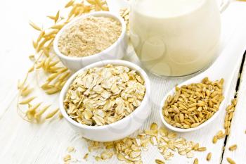Oat flakes and flour in bowls, grain in a spoon, oatmeal milk in a glass jug and ripe oaten stalks on the background of a light wooden board