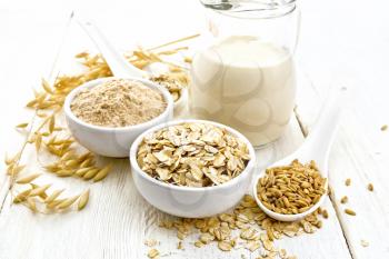 Oat flakes and flour in bowls, grain in a spoon, oatmeal milk in a glass jug and oaten stalks against a light wooden board