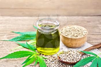 Hemp oil in a glass jar, grain in a spoon and flour in a bowl on a napkin of burlap, cannabis leaves on a wooden board background