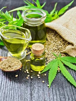 Hemp oil in two glass jars and sauceboat with grain in the bag, leaves and stalks of cannabis, a spoon with flour on the background of black wooden boards