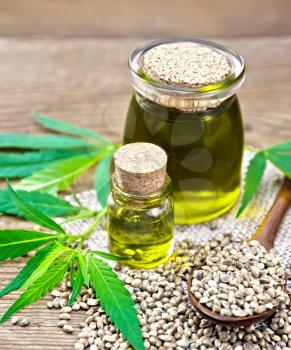 Hemp oil in two glass jars, a spoon with grains on sackcloth, leaves and stalks of cannabis on a wooden board background