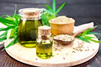 Hemp oil in two glass jars, grain in a spoon, flour in a bowl, leaves and stalks of cannabis, a napkin of burlap on a dark wooden board background
