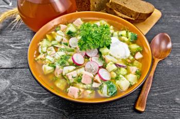 Cold soup okroshka from sausage, potatoes, eggs, radish, cucumber, greens and kvass in a bowl, bread and jug with drink on the background of wooden board