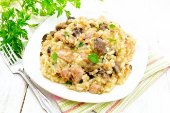 Rice risotto with mushrooms, chicken meat, cheese and garlic in a plate on towel, fork and parsley on the background of light wooden board