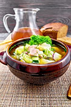Cold soup okroshka from sausage, potatoes, eggs, radishes, cucumber, greens and drink of kvass in a clay bowl, bread on wicker napkin on wooden board background