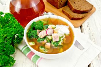 Cold soup okroshka from sausage, potatoes, eggs, radish, cucumber, greens and kvass in a white bowl on napkin, bread and jug with drink on the background of light wooden board