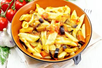 Penne pasta with eggplant and tomatoes in a bowl on napkin, fork, garlic and parsley on a wooden plank background