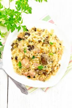 Rice risotto with mushrooms, chicken meat, cheese and garlic in a plate on towel, fork and parsley on the background of light wooden board on top