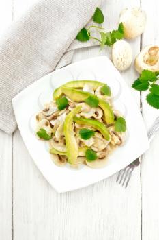 Salad from avocado and raw champignons, seasoned with lemon juice and vegetable oil with mint leaves, napkin and fork on a wooden board background from above
