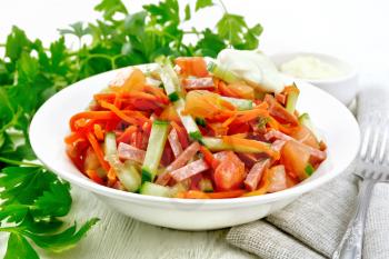 Salad from smoked sausage, spicy carrot, tomato, cucumber and spices with mayonnaise, kitchen towel, fork and parsley on a wooden board background