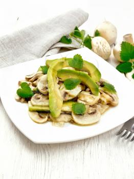 Salad from avocado and raw champignons, seasoned with lemon juice and vegetable oil with mint leaves, kitchen towel and fork on wooden board background
