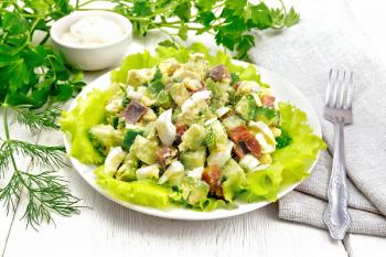 Salad from salmon, cucumber, eggs and avocado with mayonnaise on lettuce leaves in a plate, towel, dill, parsley and fork on a white wooden board background
