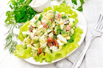 Salad from salmon, cucumber, eggs and avocado with mayonnaise on lettuce leaves in a plate, napkin, dill, parsley and fork on a light wooden board background