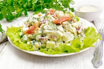 Salad from salmon, cucumber, eggs and avocado, dressed with mayonnaise on lettuce leaves in a plate, towel, dill, parsley and fork on a light wooden board background