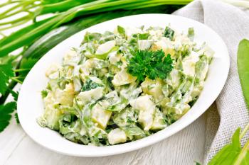 Salad of cucumber, sorrel, boiled potatoes, eggs and herbs, dressed with mayonnaise in white plate, parsley, green onions and napkin against the background of light wooden board