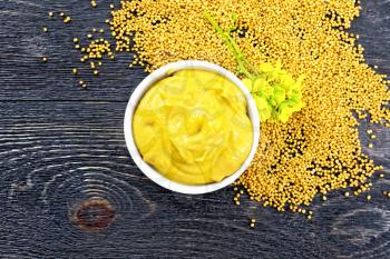 Mustard sauce in a white bowl, mustard flower and seeds on a wooden board background on top