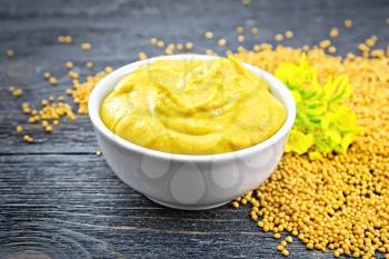 Mustard sauce in a white bowl, mustard flower and seeds on a black wooden board background