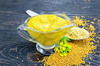 Mustard sauce in a glass sauceboat, seeds, flowers and mustard powder in a spoon on a wooden board background