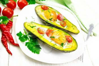 Scrambled eggs with cherry tomatoes in two halves of avocado in a plate, towel and fork on white wooden board background