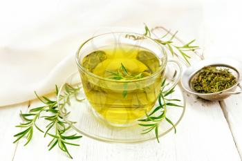 Rosemary herbal tea in a cup, a strainer with dry herb and a linen napkin on the background of a light wooden board