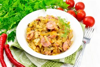 Cabbage stew with sausages in a white plate on a kitchen towel, tomatoes, parsley and a fork on the background of light wooden board