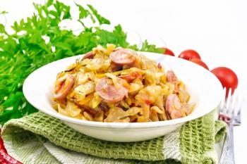 Cabbage stew with sausages in a white plate on a towel, tomatoes, parsley and a fork on the background of wooden boards