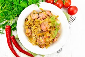 Cabbage stew with sausages in a white plate on a towel, tomatoes, parsley and a fork on the background of wooden boards from above
