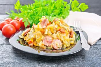 Cabbage stew with sausages in a black plate, napkin, tomatoes, parsley and fork on a wooden board background