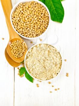 Soy flour and soybeans in two bowls, spoons and green leaves against a white wooden board from above
