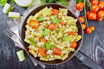 Fusilli with chicken, zucchini and tomatoes in a frying pan on burlap, forks, basil and parsley on black wooden board background from above