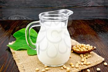 Soy milk in a jug, fresh green leaf, soybeans in a spoon and burlap on the background of a dark wooden board
