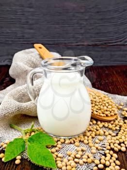 Soy milk in a jug, fresh green leaf, soybeans in a spoon and burlap on a wooden board background