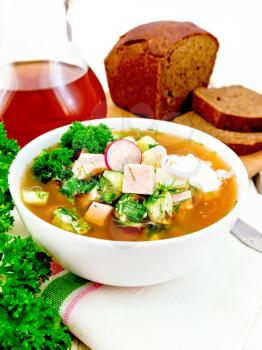 Cold soup okroshka from sausage, potato, egg, radish, cucumber, greens and kvass in a white bowl on towel, bread and jug with drink on wooden board background