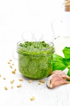 Pesto sauce in a glass jar, pine nuts, garlic, green basil and olive oil in a carafe on a background of white wooden board
