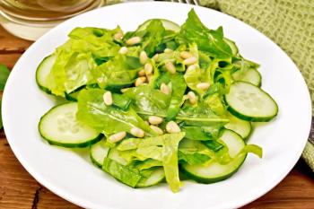 Salad from spinach, fresh cucumbers, rukkola salad, cedar nuts and spring onions, seasoned with vegetable oil on a plate, napkin and fork on a wooden board background
