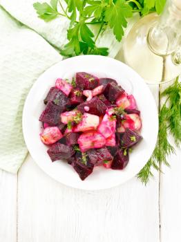 Beetroot and potato salad, seasoned with vegetable oil and vinegar in a plate, napkin, parsley and dill on the background of a light wooden board from above