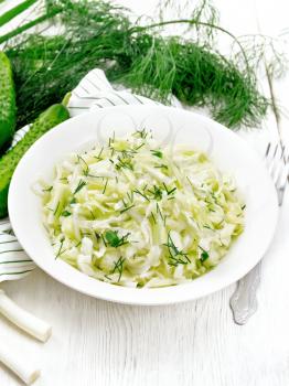 Salad of fresh cabbage, green onions and cucumber with vinegar and vegetable oil dressing in a plate, towel, dill and fork on wooden board background