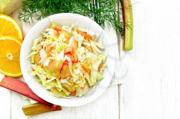 Fresh cabbage, carrot and rhubarb salad with orange juice, honey and mayonnaise dressing in a plate on a napkin, dill and fork on wooden board background from above
