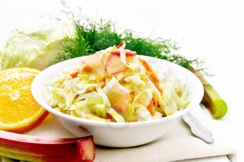 Fresh cabbage, carrot and rhubarb salad with orange juice, honey and mayonnaise dressing in a plate on a napkin, dill and fork on light wooden board background