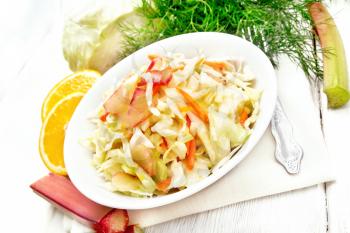 Fresh cabbage, carrot and rhubarb salad with orange juice, honey and mayonnaise dressing in a plate on a towel, dill and fork on light wooden board background
