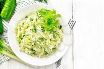 Salad of fresh cabbage, green onions and cucumber with vinegar and vegetable oil dressing in a plate, napkin, dill and fork on wooden board background from above