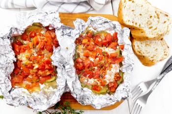 Pink salmon with zucchini, tomatoes, onions, garlic and thyme, baked in foil, fork and bread on the background of a light wooden board from above