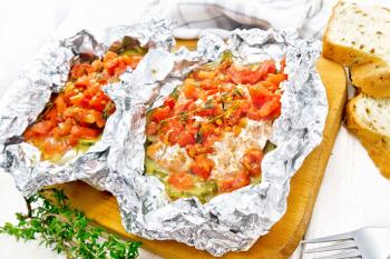 Pink salmon with zucchini, tomatoes, onions, garlic and thyme, baked in foil, fork and bread on a white wooden board background
