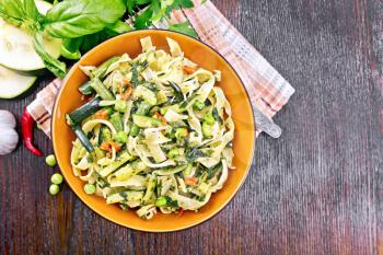 Tagliatelle pasta with zucchini, green peas, asparagus beans, hot pepper and spinach in a plate on napkin, garlic, fork and basil on wooden board background from above