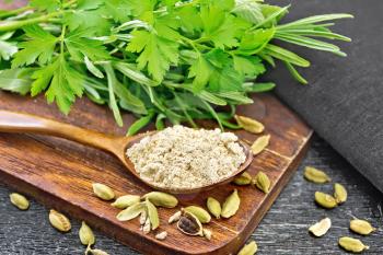 Ground cardamom in a spoon, seasoning capsules, napkin, fresh parsley and rosemary on wooden board background